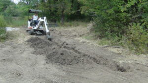 Individual on a bob cat, small digging device, digging a drainage trench in the exposed dirt. 