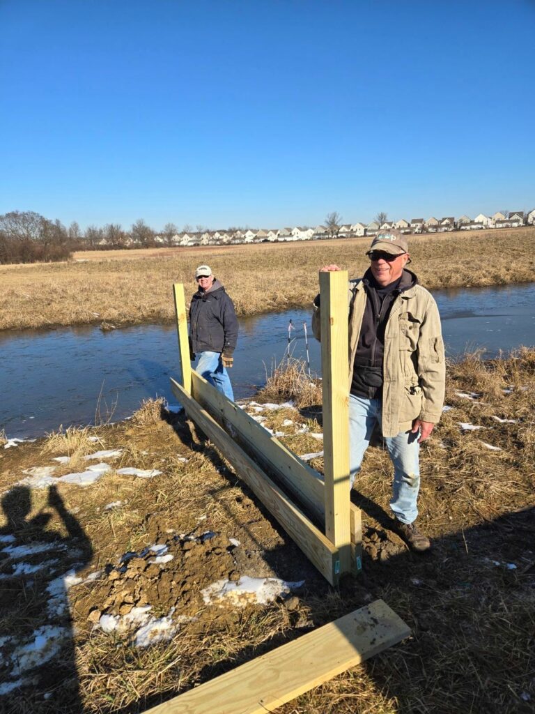 Two volunteers stand at the corners of the rectangle framing that will be our new wetland deck, They are helping to brace the corner posts of the platform.